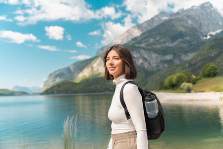 Wandering off the grid, a smiling woman walks a winding path through lush greenery, with mountain peaks and a calm lake setting the tone for mindful summer travelの写真素材