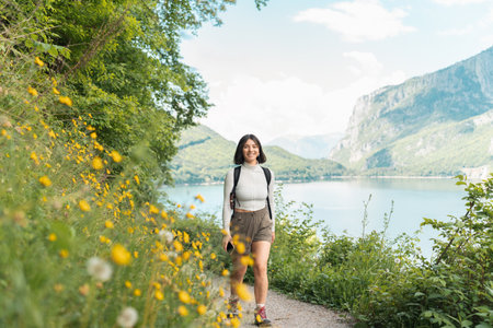 Glowing with joy, a woman strolls a nature trail wrapped in greenery, grinning as mountains and a sparkling lake unfold her perfect summer resetの写真素材