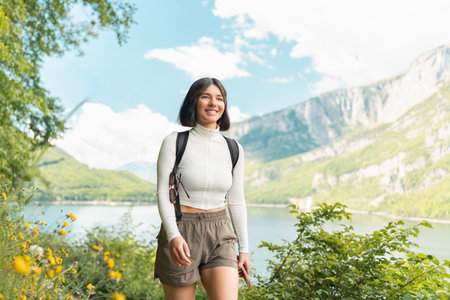 Smiling wide, a woman follows a peaceful trail through alpine greenery, with lake reflections and sun-kissed peaks echoing a laid-back summer escapeの写真素材