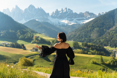 A stylish young woman in a black dress walks through sunlit fields at sunrise, with the Dolomites rising behind her. A dreamy escape into nature and eleganceの写真素材