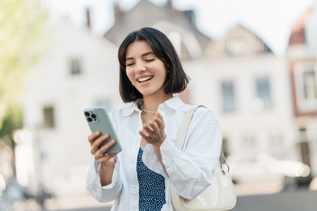 Quick check. Middle Eastern woman with short hair and a calm smile manages finances on her phone while standing on the sidewalk in a white shirtの写真素材