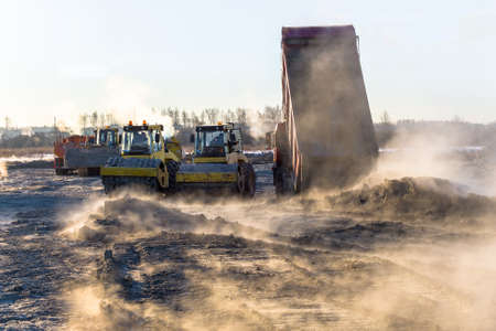 roller compacts soil in embankment on the road's construction. sand consolidation on road-building. Compactor driving on sandy. wheel marks on the sand. hard work. transportation of sand by trucksの写真素材