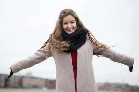 Smiling young girl in a coat spread his arms. Girl in a black coat, a scarf and a red dress against a gray sky. Not Isolated. Autumn bank overcastの写真素材