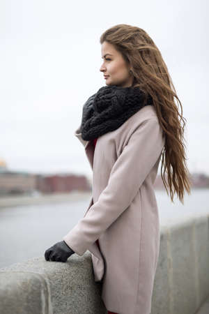 Mysterious young woman stands on the embankment and looking into the distance. Girl in a black coat, a scarf and a red dress against a gray sky. Not Isolated. Autumn bank overcastの写真素材
