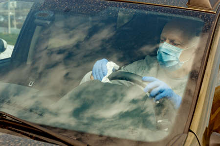 Car disinfection. A man in gloves and a mask wipes the steering wheel with a disinfectant wipe.の写真素材