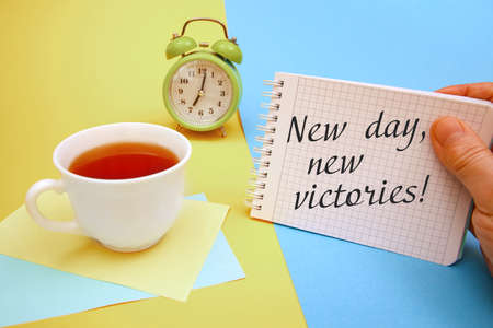 Cup of tea and a notebook with the inscription New day, new victories. Motivation of a good mood.