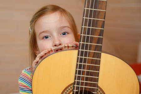 A little girl peeks out from behind a guitar case at home.