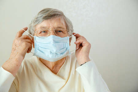 An elderly woman in glasses puts on a medical mask on her face. Elderly woman in glasses with a medical mask worries about coronavirusの写真素材