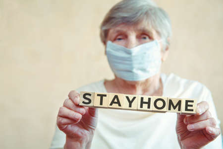 An elderly woman with a medical mask on her face holds cubes with the inscription STAY HOME. The concept - quarantine, isolation, caronovirus Covid-19の写真素材