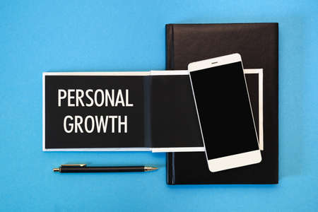 A white notebook with black pages, a smartphone and a pen on a blue background. The inscription PERSONAL GROWTH on the notepad.の写真素材