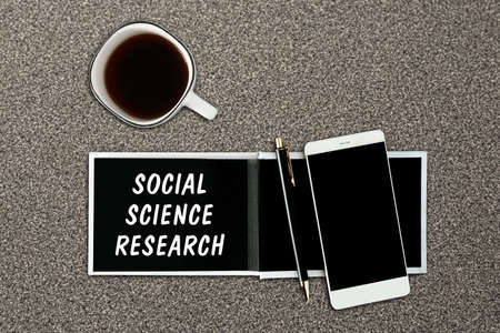 A notebook with black pages, smartphone and a cup of coffee on a gray background. The inscription Social Science Research on the notepad.の写真素材