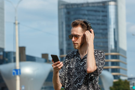 A young man with a smartphone in headphones and sunglasses listens to music on the background of buildings modernの写真素材