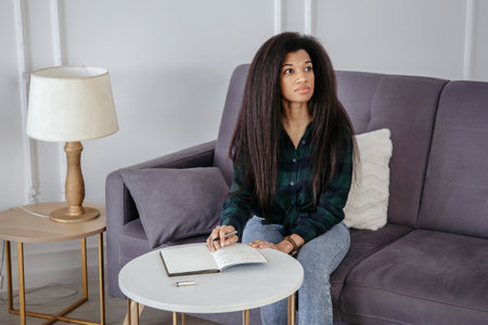 A beautiful African-American girl with a notebook in her hands sits on the sofa and makes notes in a notebook.の写真素材