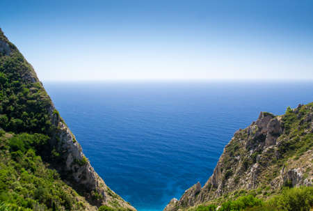 Rocky cliffs with green vegetation in foreground and blue sea in background on a sunny dayの写真素材