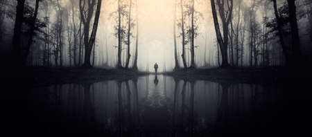 Surreal symmetrical lake in dark haunted Halloween forest with silhouette of manの写真素材