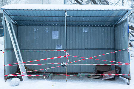 metal shed for garbage containers closed to prohibit the passage of ribbons, red and white color, winter is snow, bags of garbage and discarded interior doors with glass, crumpled cardboard boxes and ads on the wall of the shedの写真素材