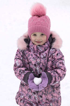 Beautiful smiling girl in winter in mittens holding snow in the shape of a heart. The street is snow and very cold. The girl festive mood. She gives you a gift.の写真素材