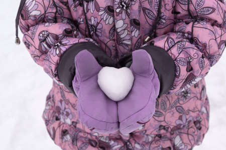 Girl in winter pink jacket and purple mittens holding a snowball in the shape of a heart. It's snowing and very cold. She gives you a gift.の写真素材