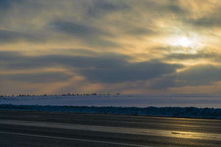 Winter. In the foreground a country paved two lane road. In the background field covered in white snow. The distance the planting of trees. In the sky crimson clouds. Through the clouds and the shining yellow sun.の写真素材