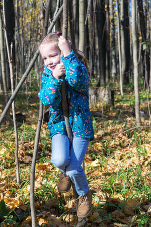 Little girl climbs a tree in the woods or Park. Autumn, Sunny day, the earth growing grass and fallen leaves are different colors. The girl is dressed in a jacket, jeans and boots.の写真素材
