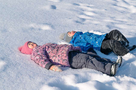 Children, brother and sister lying in the snow on the back. Laugh and smile. Dressed in jackets, hats, warm pants, boots, boots and mittens. Wear pink, blue, grey and black. He closed his eyes.の写真素材