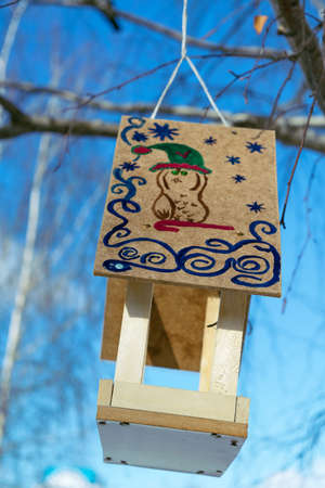 A bird feeder with a roof. On the roof of a drawn figure. The animal depicted in the cap with a curved stick, around patterns and snowflakes. Feeder hanging on a tree on a rope. It is made of wood.の写真素材