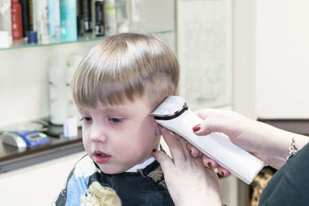 Little cute boy sitting in the chair at the hair salon. Covered with a cloak. It cut Barber electric hair clippers white. In the background are accessories.の写真素材