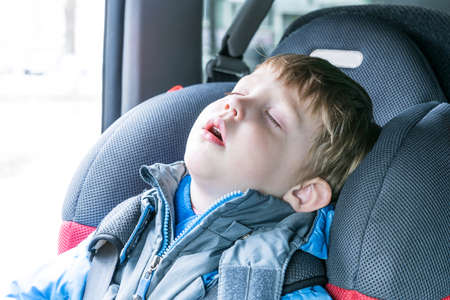 Little boy sleeping in child car seat. The chair is very comfortable. A boy wearing a blue jacket and gray. Boy wearing safety belts. He closed his eyes and opened his mouth.の写真素材