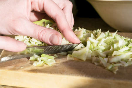 A woman with a knife cut fresh cabbage on a cutting wooden Board. One hand holds a knife, the other hand holds the cabbage. In the background is a plate.の写真素材