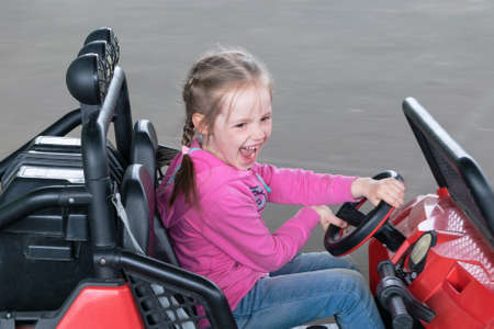 Little girl rides on toy electric cars in the Playground. The child is very happy, has fun and entertains. His mouth opened with joy. Girl rides the speed and is behind the wheel.の写真素材