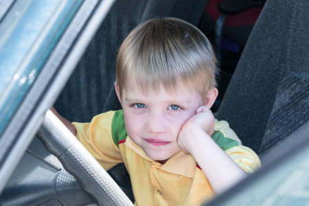 Little boy sitting in the car driving. One hand props her cheek. The other hand held behind the wheel and looking at us. He's thoughtful and smart look. He dreams about something.の写真素材