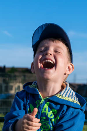 A fun, happy little boy laughs very much. The boy closed his eyes in pleasure and opened your mouth wide while laughing. In the background the blue sky and building.の写真素材