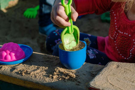 Little girl playing in a sandbox with a plastic shovel and a mug. In a mug filled with the sand, lie next to a plastic plate and the mold. Summer, day, sun shining.の写真素材