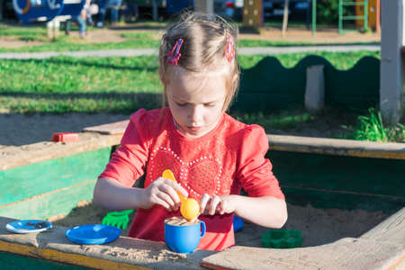 Little girl playing in a sandbox with a plastic shovel and a mug. In a mug filled with the sand, lie next to a plastic plate and the mold. Summer, day, sun shining.の写真素材