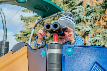 Little boy looking through binoculars on a slide at the Playground. The boy is holding the binoculars with your hands and looking for something in the distance. He is interested and very intelligent.の写真素材