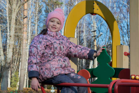 Little girl sitting on the slide at the Playground and smiles. Looking at us and holding his hand over the wooden fir. Day, the sun shining in the background the trees in the Park. The girl is happy.の写真素材