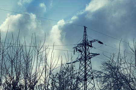 Support or tower power lines against the dark sky and tree branches. Rainy clouds, cloudy. Threat of termination of power supply. Frozen tree branches in the foreground.の写真素材