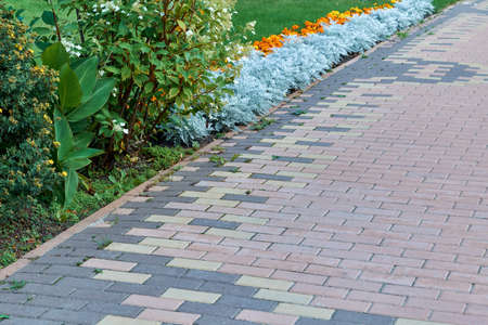 Paving pattern with rectangular shape and brown color. Along paved walkways planted flowerbed with beautiful orange and yellow flowers. In the background, lawn with green grass.の写真素材
