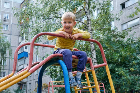 The boy is sitting on a children's slide and looks at us joyfully. Useful rest on the Playground. Outside in the fresh air. Physical activity for health. The hill is iron and painted with paint.の写真素材