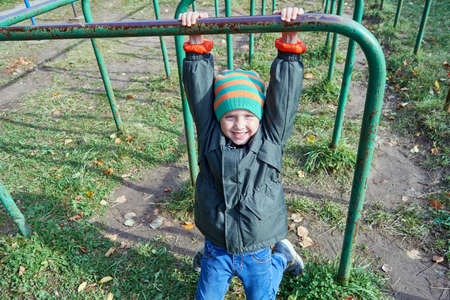 A little boy is hanging on the bar, looking at us and smiling. Exercise in the fresh air. Useful education for health. Development of physical strength, dexterity, endurance.の写真素材