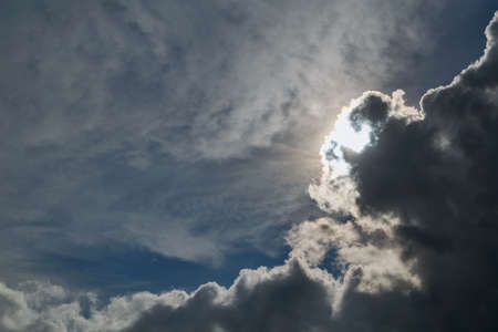 The sun shines brightly in the clear blue sky. Big storm clouds are coming. Cumulus clouds before rain looming on the sun and want his shield. Sunlight and rays illuminate the sky and clouds.の写真素材