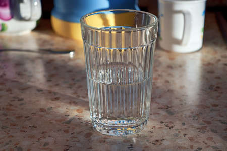 On the kitchen table there was a glass of water is half full. The faceted glass. Kitchen utensils are visible in the background. Sunlight illuminates the table. Clean water is good for your health.の写真素材