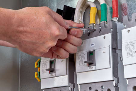 A person or worker screws an electric circuit breaker with a key to the circuit Board in the electrical Cabinet. Three powerful switches are fixed in a row. Switches are connected to cables or wires.の写真素材