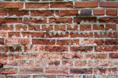 Old red brick wall of rectangular shape with triangular cutouts. Pieces of bricks partially broke off and cracked. Some bricks are of non-standard shape with cutouts of wavy shape.の写真素材
