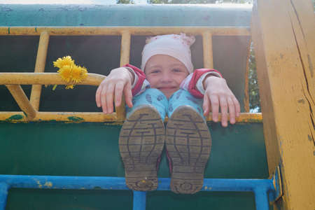 The girl jokes and plays at the children's sports complex. She sticks out her arms and legs and face and smiles. Not see the body. Good mood. Dandelions lie on the crossbar. Bright sunny day.の写真素材