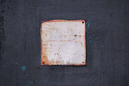 A square aluminum sign with a red frame on an old painted metal black wall. The sign is rusted and painted white. The paint on the black wall is cracked and peeling. Industrial background, backdrop.の写真素材