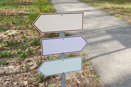 A road sign with three arrow-shaped signs pointing in different directions. Pointers are empty. In the background there is an asphalt path, earth and grass. The signs have colored borders.の写真素材