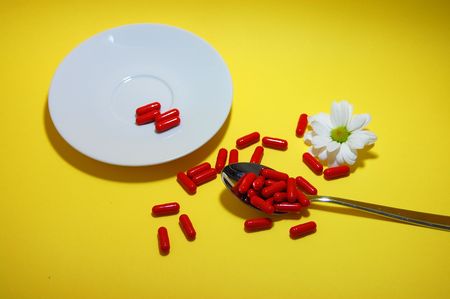 Red pills scattered over the white plate and a spoon with a daisy on a yellow backgroundの写真素材