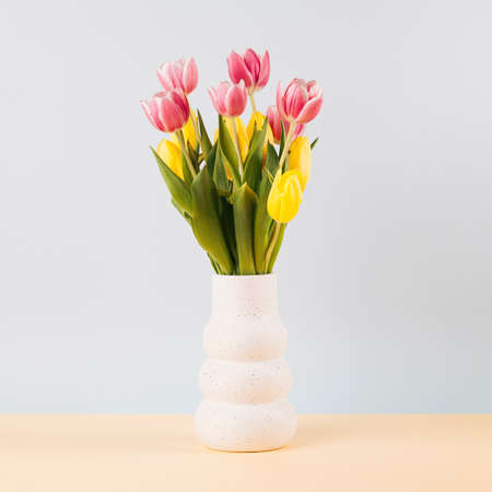 Bouquet of yellow and pink tulip flowers in transparent glass vase on sand coloured table. Simple composition on light pastel blue background.の写真素材