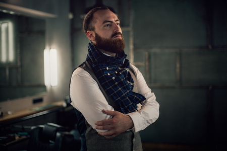Handsome bearded man hipster with stylish beard in white shirt and chekered scarf standing with serious face in beauty salon or barbershopの写真素材